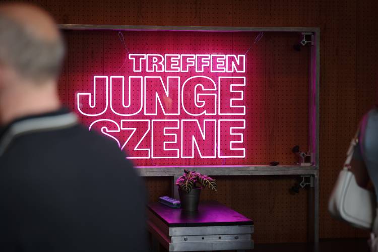 An der Wand ist ein leuchtender, pinkfarbener Schriftzug mit der Aufschrift „Treffen junge Szene“ zu sehen, der in dem ansonsten dunklen Raum besonders hervorsticht.