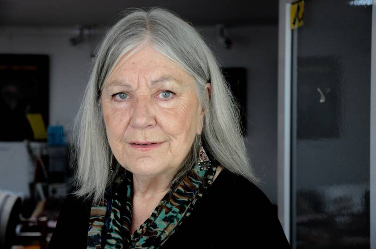 Helga Schubert – Ältere Person mit langen grauen Haaren und Ohrringen, trägt einen gemusterten Schal vor grauem Hintergrund.