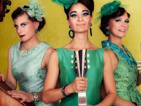 Zucchini Sistaz „Eleganz oder gar nicht“ – Drei Personen in grünen Kleidern mit Instrumenten: Kontrabass, Mandoline, Trompete. Jede trägt grünen Kopfschmuck, Hintergrund gelb.