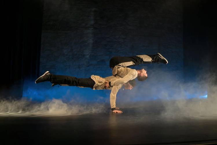 George Kranz & friends – Zwei Personen führen eine Handstand-Akrobatik aus. Eine Person balanciert waagerecht auf dem Rücken der anderen in einem Bühnennebel.