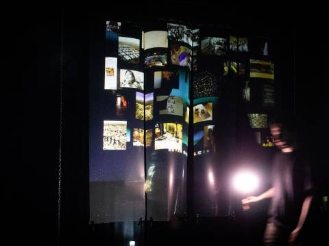 Mei Liu, Homesick for Another World (2025). Foto: Jia Y. Ding – Beleuchtete Projektionsflaeche mit vielen Fotos in Reihen. Unscharfe Person rechts haelt eine Lampe. Umgebung ist dunkel.