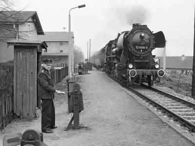 Ein Schrankenwärter steht vor einer rekonstruierten Kriegslok der Baureihe 52. – Das Schwarzweißfoto zeigt links einen Schrankenwärter, der am Gleis auf der rechten Seite vor einer rekonstruierten Kriegslok der Baureihe 52 steht