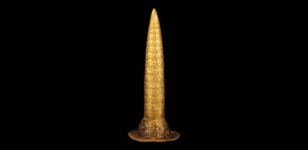 Den Berliner Goldhut zieren verschiedene Kreisornamente, die als lunisolarer, d.h. auf Sonne und Mond basierenden Kalender verstanden werden können. Sie stehen für die Verbindung von Kult, Wissen und auch materiellem Reichtum. – Goldener, verzierter konischer Helm auf schwarzem Hintergrund. Kunstwerk mit kreisförmigen Mustern in parallel verlaufenden Reihen.