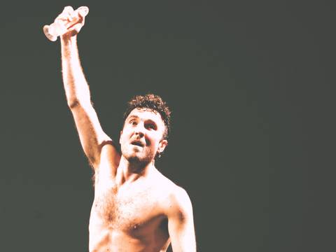  – Performer mit nacktem Oberkörper auf dunkler Bühne, Arm erhoben, transparenter Dildo in der Hand.