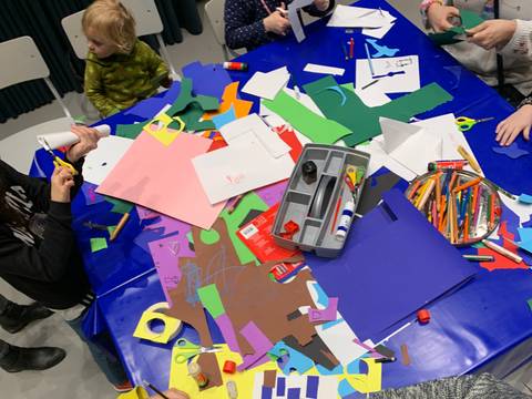 © Nancy Jesse – Man sieht viele Kinder an einem großen Basteltisch sitzen. Auf dem Tisch liegen viele bunte Bastelaccessoires.