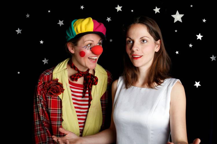 Zwei Personen vor schwarzem Hintergrund mit Sternen. Links Clown mit rotem Hut, roter Nase und kariertem Mantel. Rechts Person im weissen Kleid, neutraler