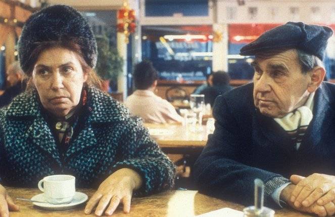 Zwei Personen sitzen an einem Tisch in einem Cafe. Links eine weiblich gelesene Person mit Fellhut, rechts ein maennlich gelesener Mann mit Schiebermuetze.