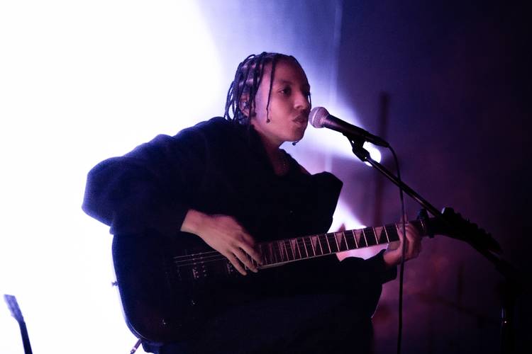 Person mit geflochtenem Haar spielt Gitarre und singt in ein Mikrofon vor hellem Hintergrund.