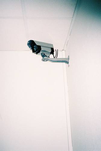 © Murat Aslan – Überwachungskamera an der Ecke einer Raumdecke, montiert auf einem Metallarm. Wände und Decke sind weiß.
