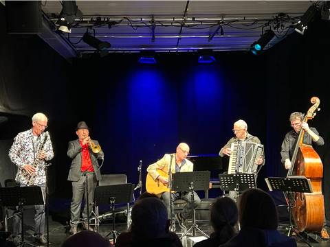  – Fünf Musiker auf einer Bühne: Saxophon, Trompete, Gitarre, Akkordeon, Kontrabass. Publikum im Vordergrund, blauer Lichthintergrund.