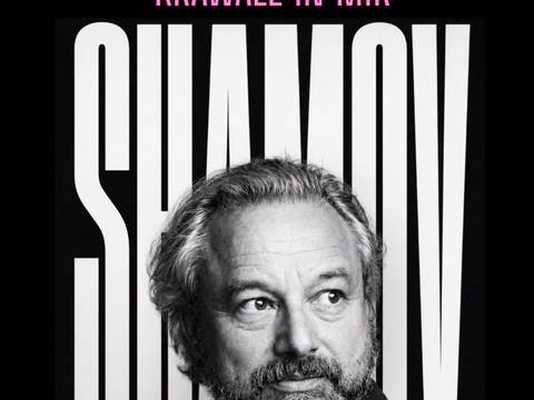  – Schwarzweiß-Porträt von Roman Shamov mit mittellangem, grauem Haar und Vollbart. Roman Shamov blickt leicht nach rechts. Im Hintergrund befinden sich vertikale schwarze Streifen auf hellem Grund. Die Aufnahme zeigt sein Gesicht und den oberen Schulterbereich und wirkt kontrastreich sowie professionell fotografiert.