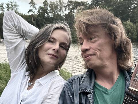  – Kerstin Reimann und Karsten Troyke sitzen lächelnd nebeneinander am Ufer eines Sees. Kerstin Reimann links trägt eine weiße Bluse und hat einen Arm hinter den Kopf gelegt, Karsten Troyke rechts hält eine Gitarre und blickt sie mit einem warmen Lächeln an. Im Hintergrund sind Wasser, Schilf und ein bewaldetes Ufer zu sehen.