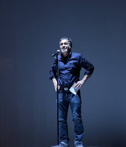 Jürgen Kuttner, Foto: Thomas Aurin – Person steht mit Mikrofon auf Bühne, trägt dunkles Hemd und Jeans, Papier in der linken Hand, neutrale Mimik, dunkler Hintergrund.