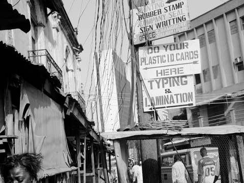 Akinbode Akinbiyi, Lagos Island, Lagos, 2004, from the series: "Lagos: All Roads", since the 1980s – Quadratische Schwarz-Weiß-Fotografie: Blick auf verschiedene Häuserfassaden, zwischen denen eine Straße entlangführt neben der Personen laufen. In der Bildmitte ist ein Strommast zu sehen, an den Reklametafeln mit Aufschriften wie „Typing & Lamination“ angebracht sind. Im vorderen, linken Teil der Fotografie ist eine weiblich-gelesene Person mit weit geöffnetem Mund und nach unten gesenktem Blick zu sehen.