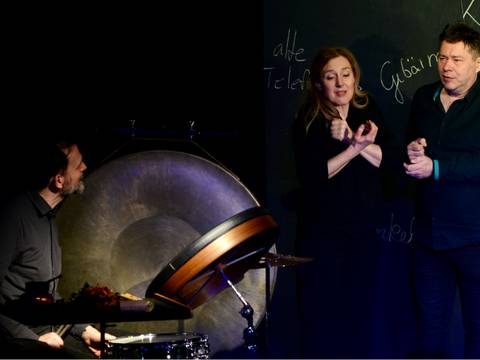  – Drei Personen, eine mit Drum und Gong, zwei gestikulierend vor Tafel mit den Worten alte, Tele, Geburt, Küche.