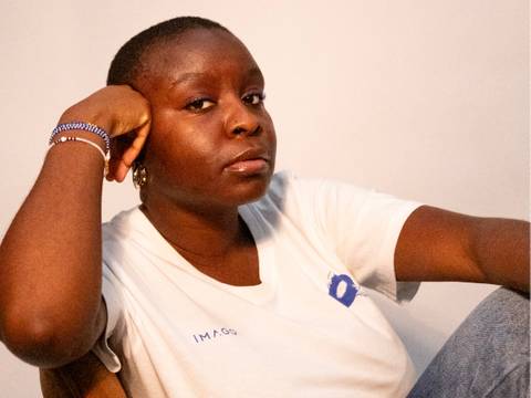  – Person in weißem T-Shirt mit Aufschrift "IMAGO", sitzt, stützt den Kopf auf der Hand, schaut ernst, trägt Armbänder und Ohrringe.