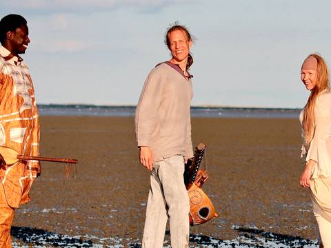  – Drei Personen stehen am Strand, jede hält ein Saiteninstrument, lächelnd, mit dem Meer im Hintergrund.