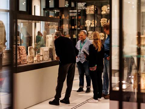Besucher*innen einer Führung im Bereich "Grafische Kommunikationssysteme in Mesoamerika" des Ethnologischen Museums – Personengruppe betrachtet antike Artefakte in einer modernen Museumsvitrine. Im Hintergrund weitere Ausstellungsstücke hinter Glas.