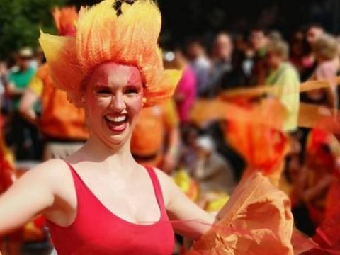 Feuerfunken und Regenglück - nach dem Märchen der Regentrude – Person in rotem Oberteil mit orange-gelber Kopfbedeckung, gelber und roter Stoff, lachend, dicht gedrängte Menschenmenge im Hintergrund.