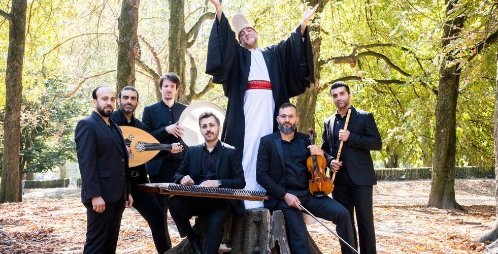 Sieben Personen mit Musikinstrumenten im Wald. Eine davon trägt ein weißes Gewand mit rotem Gürtel und Cap. Die anderen tragen schwarze Anzüge.