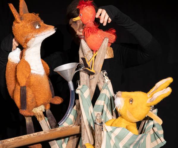 Der Koffer – Ein Fuchs, ein roter Vogel und ein gelber Hase als Puppen; eine Person in Schwarz im Hintergrund steuert die Figuren.