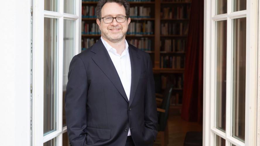 Eine Person mit Brille und Anzug steht lächelnd in einer Türöffnung; im Hintergrund ein Bücherregal.