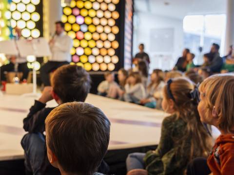  – Eine Gruppe von Kindern sitzt auf dem Boden vor einer Bühne, auf der zwei Personen musizieren. Im Hintergrund leuchtet eine bunte Wabenwand.