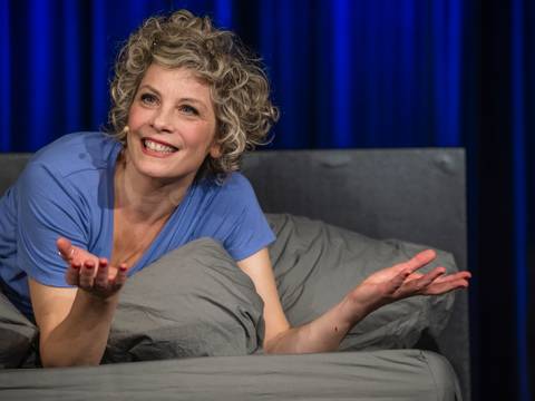  – Person mit Locken und Mikrofon liegt auf grauem Bett, trägt blaues T-Shirt, breitet lachend die Arme aus, blauer Hintergrund.