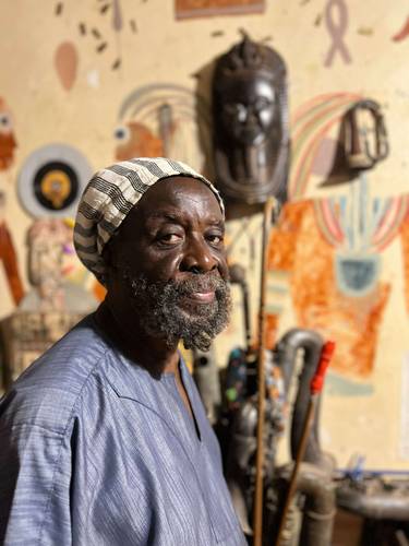 Nii Noi Nortey. Foto: Ato Annan – Person mit gestreiftem Hut und blauem Gewand, lächelnd vor kunstvoll bemalter Wand und Skulpturen.