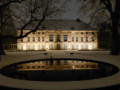 Schloss Schönhausen im Winter – Ein großes, nachts beleuchtetes Gebäude mit symmetrischer Fassade und hohen Bogenfenstern, umgeben von kahlen Bäumen, deren Spiegelbild sich in einem runden Teich im Vordergrund widerspiegelt: Schloss Schönhausen im Winter