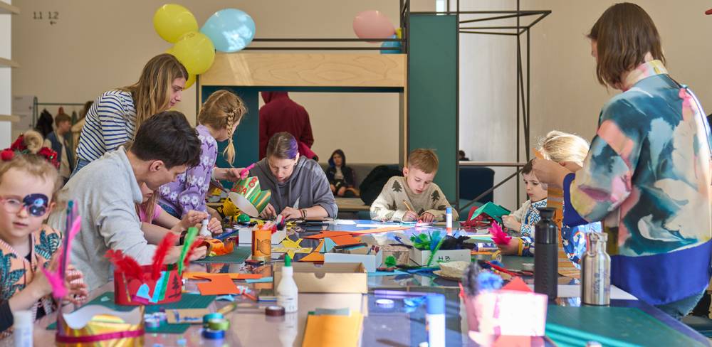 Kinder und Erwachsene basteln am Tisch mit buntem Papier und Federn. Auf dem Tisch stehen Bastelmaterialien. Im Hintergrund hängen bunte Luftballons.