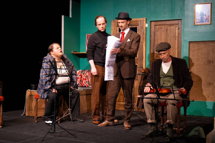 Vier Personen stehen auf einer Theaterbühne. Eine Person liest ein großes Papier. Zwei Musikinstrumente stehen vor den anderen Personen.