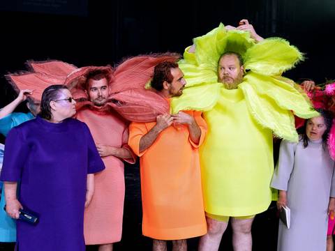 – Sechs Personen in bunten Kostuemen mit großen, blütenartigen Halskrausen. Farben sind lila, rosa, orange, neongelb, hellgrau und pink. Schwarz als Hintergrund.