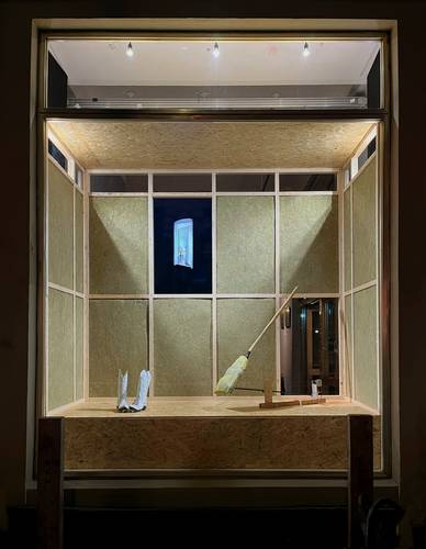 Künstlerinnengespräch »A house and two windows« – Holzrahmen mit Besen, zwei weißen Schuhen und kleinem Bildschirm in Schaufenster. Obere Beleuchtung und dunkler Hintergrund.