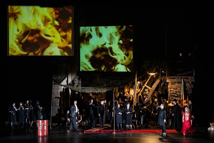 La forza del destino – Bühnenbild mit 15 Personen in schwarzer Kleidung, rote Tonne mit V, zwei Projektionen von Flammen, rechte Person in rotem Kleid.