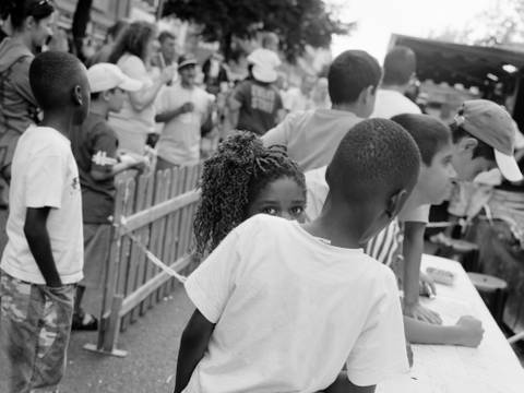 Akinbode Akinbiyi, Wedding, Berlin, 2005, Aus der Serie: „African Quarter“, seit den 1990er Jahren – Schwarz-weiß-Fotografie: Im quadratischen Format zeigt die Fotografie mehrere Personen, die vorwiegend mit dem Rücken zur Kamera stehen, während eine junge, weiblich gelesene Person direkt in die Kamera blickt.