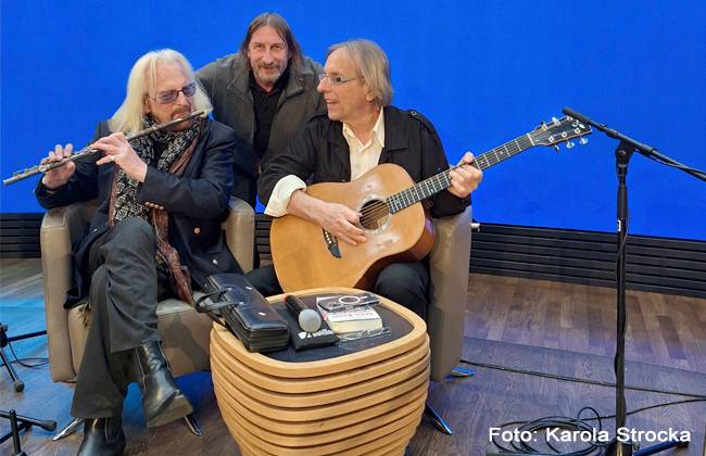 STASI-KNAST & OSTSEEFLUCHT – Drei Personen, zwei sitzen mit Gitarre und Querflöte auf Sesseln, eine steht hinten. Tisch mit Mikrofon, Buch und CD. Blauer Hintergrund, Text: Foto: Karola