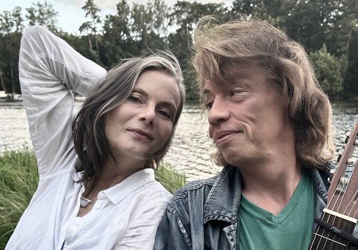 Kerstin Reimann und Karsten Troyke sitzen lächelnd nebeneinander am Ufer eines Sees. Kerstin Reimann links trägt eine weiße Bluse und hat einen Arm hinter den Kopf gelegt, Karsten Troyke rechts hält eine Gitarre und blickt sie mit einem warmen Lächeln an. Im Hintergrund sind Wasser, Schilf und ein bewaldetes Ufer zu sehen.