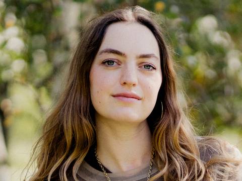 Dana Vowinckel – Person mit langen braunen Haaren und Kette steht draußen vor unscharfem Hintergrund. Text im Bild: © Max Zerrahn / Suhrkamp Verlag.
