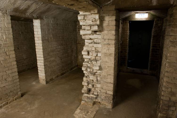 Keller der "Baracke 13" – Unterirdischer Raum mit grob verputzten Ziegelwänden, mehreren Pfeilern und einem beleuchteten Ausgangsschild über einer offenen Tür.