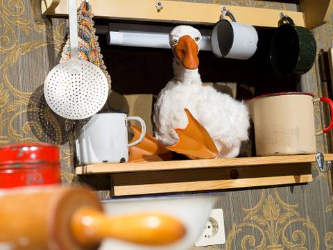  – Kuscheltier-Gans auf einem Regal zwischen Emaillebechern, daneben ein Sieb und ein Nudelholz vor einer dekorativen Tapete.