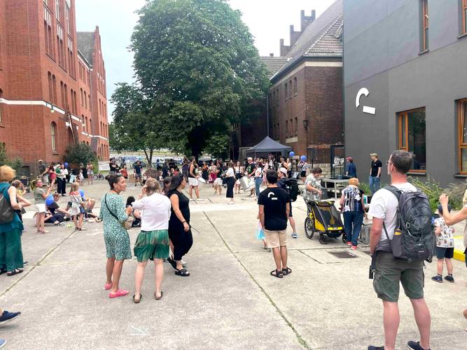 Menschen auf einem Hof mit roten Backsteingebäuden und Bäumen, ein Kind im Kinderwagen, ein Pavillon im Hintergrund, viele tragen helle Sommerkleidung.