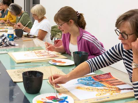  – Erwachsene Personen sitzen an einem Tisch und malen auf Papier. Sie verwenden Pinsel und Farben. Keine spezielle Bildtechnik erkennbar.