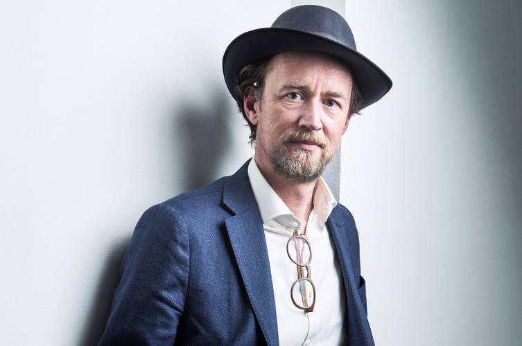 Mathias Brodkorb – Person mit Hut und Bart, trägt einen blauen Anzug und ein weißes Hemd. Brille hängt am Hemd. Blickt geradeaus, an eine weiße Wand gelehnt.