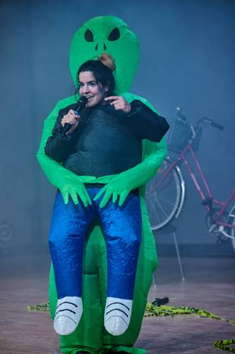 Person im Alienkostüm mit Mikrofon auf Bühne, Hintergrund mit Fahrradteil sichtbar, unscharfe Umgebung.