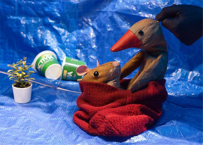 Zwei Stofftiere, Vogel und Maus, in rotem Stoff auf blauer Plane. Ein Pflanzentopf und umgefallene grüne Becher mit "Activia" im Hintergrund.