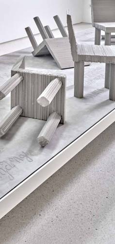 Bilddetail des Denkmals „Never Forget“ – Graue, umgestürzte Stühle und Tisch aus Wellpappe auf hellem Boden. Strukturiertes Material erkennbar. Keine weiteren Elemente sichtbar.