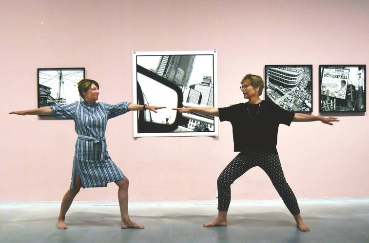 Yoga in der Ausstellung „Akinbode Akinbiyi. Being, Seeing, Wandering“ – Foto: Zwei Personen stehen sich in Yoga-Pose gegenüber und blicken einander an. Im Hintergrund sind schwarz-weiß-Fotografien von Akinbode Akinbiyi vor einer rosafarbenen Wand zu sehen.