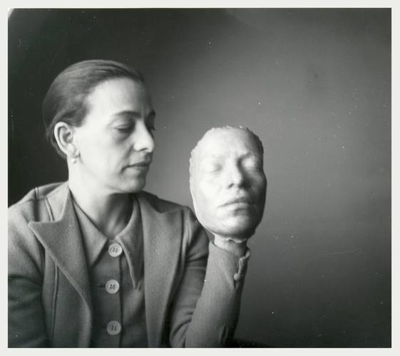 Helene Weigel - Revolutionärin im Rampenlicht – Eine Person haelt eine Maske mit geschlossenen Augen in der Hand. Sie traegt ein Jackett mit drei Knöpfen und hat glattes, zurückgebundenes Haar.