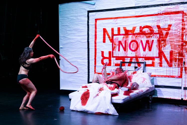 Three Times Left Is Right – Tanzende Frau in Unterwäsche mit Gedärmen und blutverschmierter Mann im Bett vor einer Plakatierung mit der Aufschrift "Now" und "Never Again".